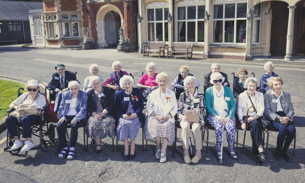 Group photo of Bletchley Park Veterans sat in two rows in front of the Bletchley Park Mansion