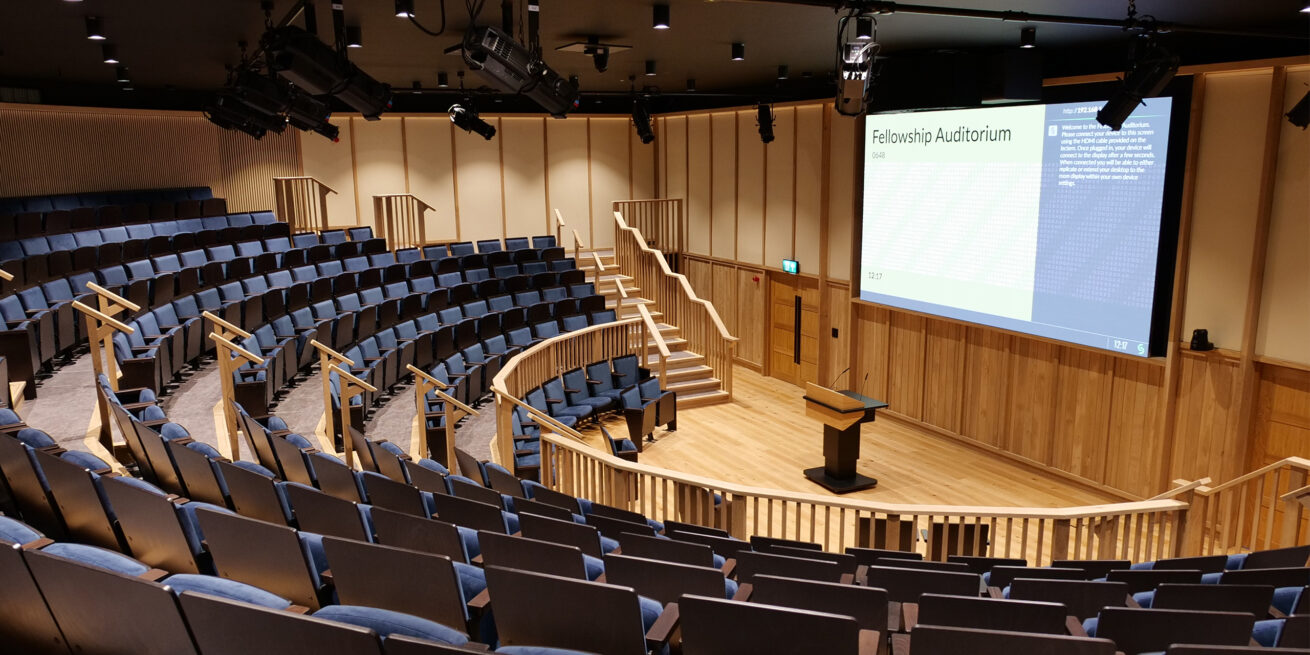 An image of inside the Fellowship Auditorium. The room has no people in it. There is a large screen on the righthand side wall. In front on the ground is a lectern. There is a semi-circular space in front of the lectern and then rows of blue seats with each row higher than the last.