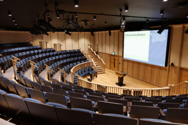 An image of inside the Fellowship Auditorium. The room has no people in it. There is a large screen on the righthand side wall. In front on the ground is a lectern. There is a semi-circular space in front of the lectern and then rows of blue seats with each row higher than the last.